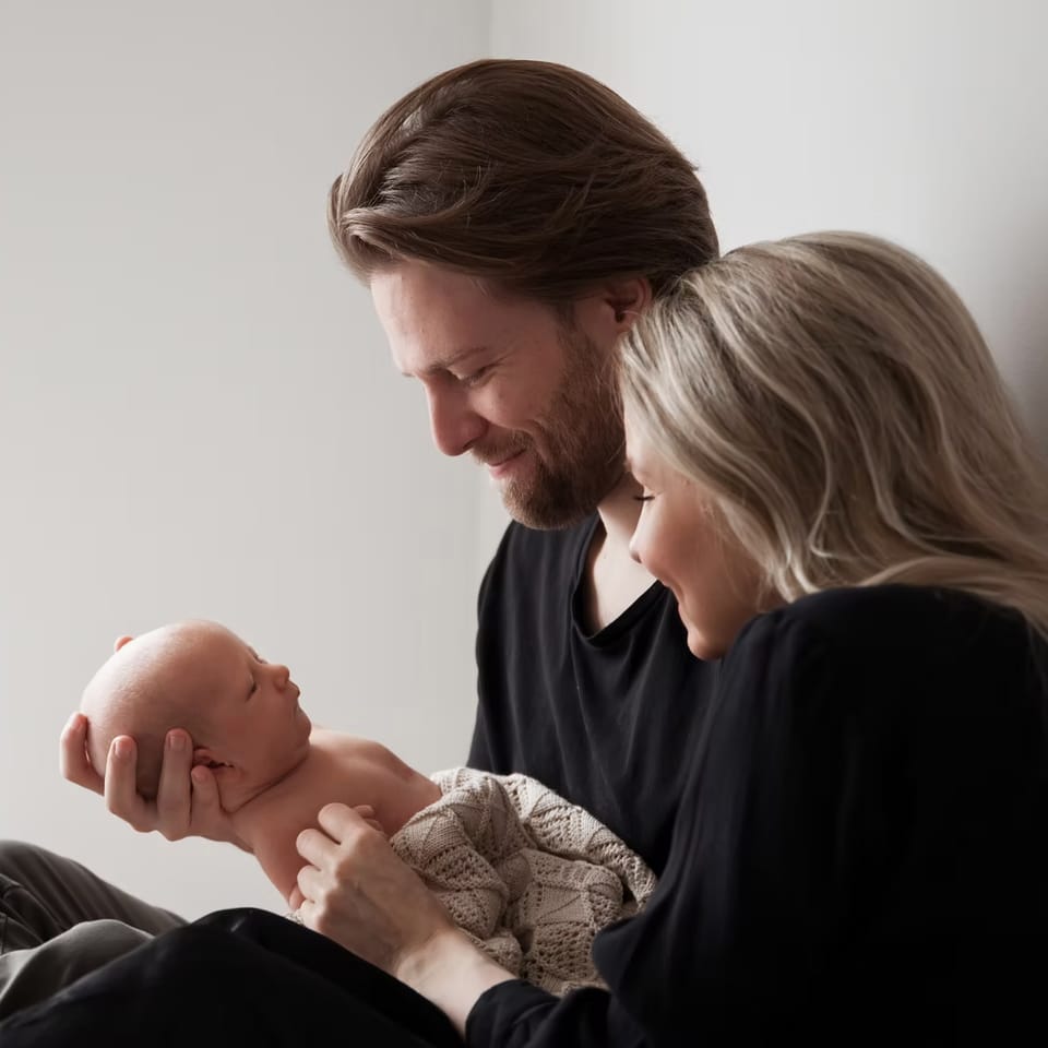 Et forældrepar sidder i hjemmet og smiler til deres nyfødte, som faren holder i hænderne